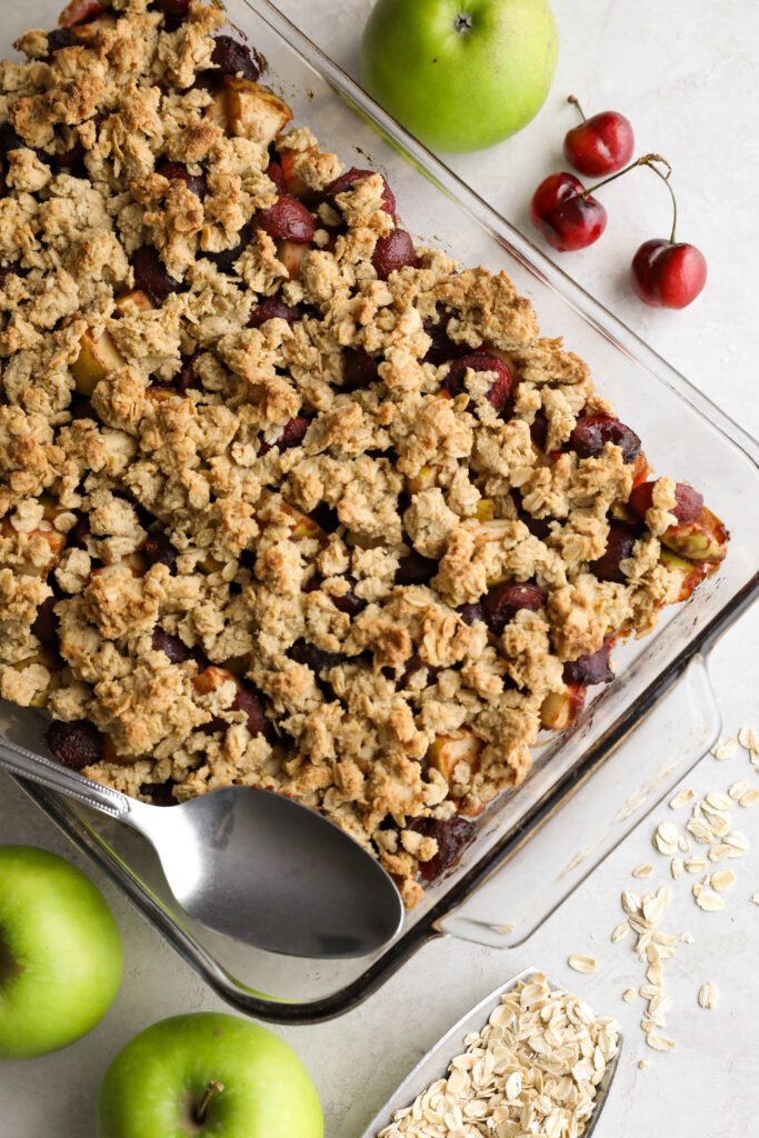 Apple Cherry Tahini Crumble baked in glass dish with a spoon surrounded by cherries and apples