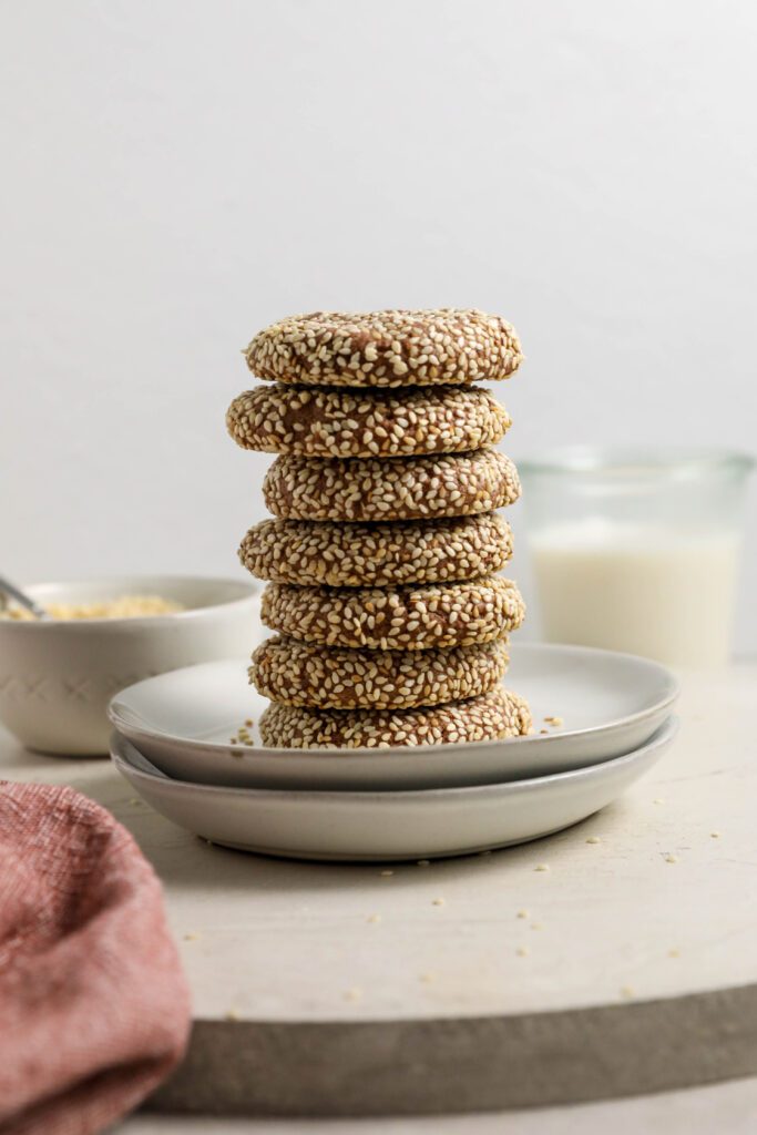 Chocolate Sesame Tahini Cookies stacked on two plates with glass of milk and bowl of sesame seeds in background by Flora & Vino