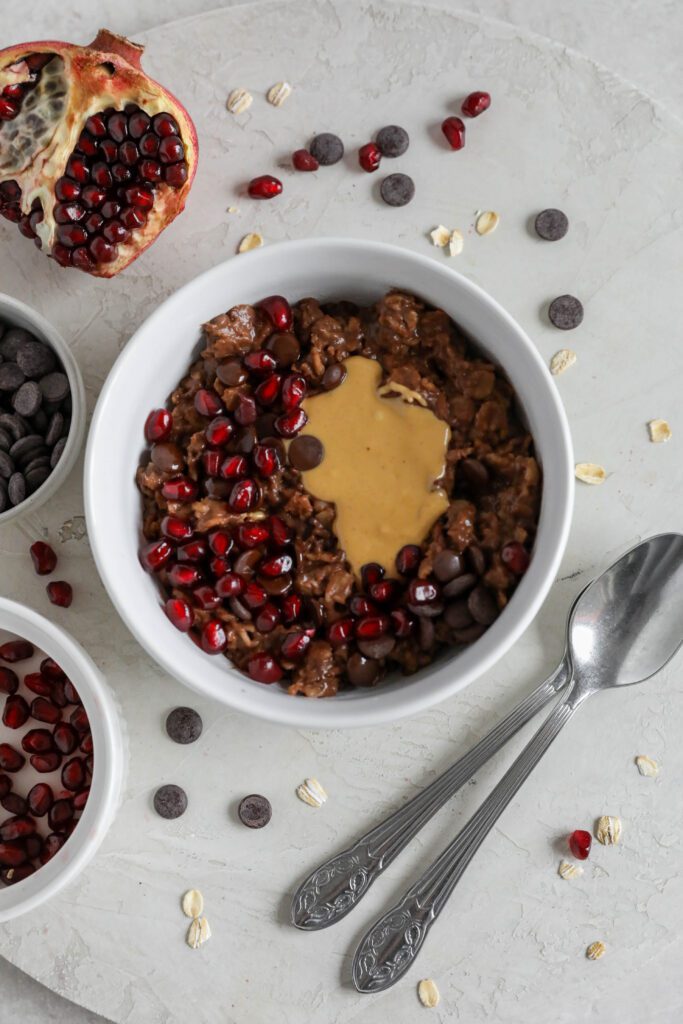Chocolate Stovetop Oats served in a bowl topped with peanut butter, dark chocolate chips, and pomegranate seeds with two spoons on the side by Flora & Vino