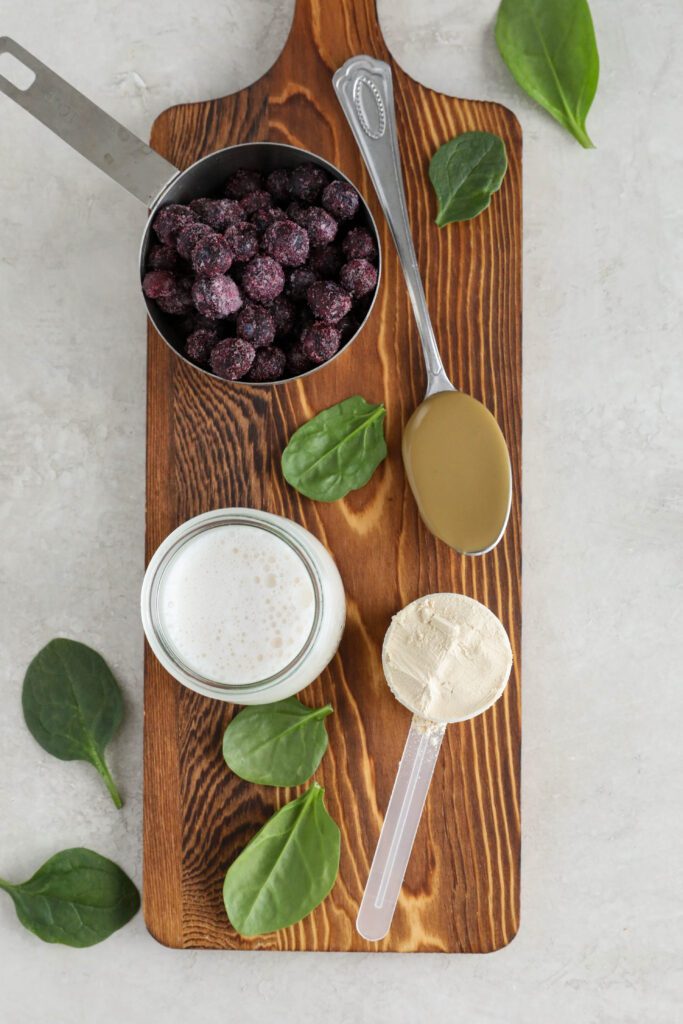 frozen blueberries, spinach, almond milk, SunButter on a spoon, and protein powder on wooden board by Flora & Vino