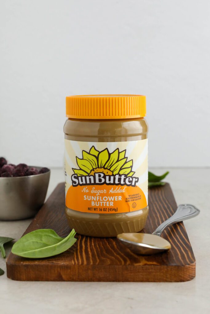 SunButter jar on wooden board with spoon next to it with spinach leaves and frozen blueberries in background by Flora & Vino