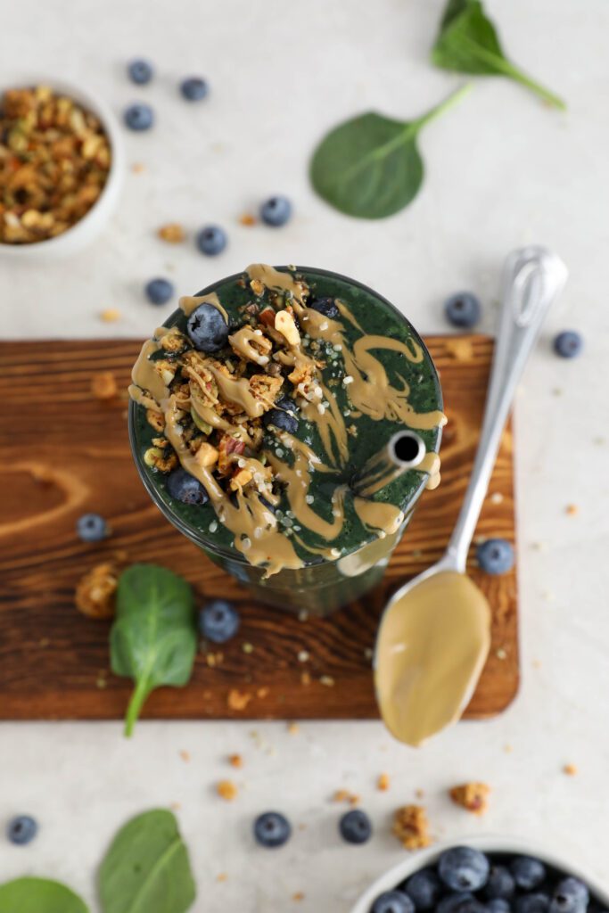 My Go-To Green Smoothie served on wooden board surrounded by blueberries and spinach leaves topped with granola with a spoon with SunButter next to the glass by Flora & Vino