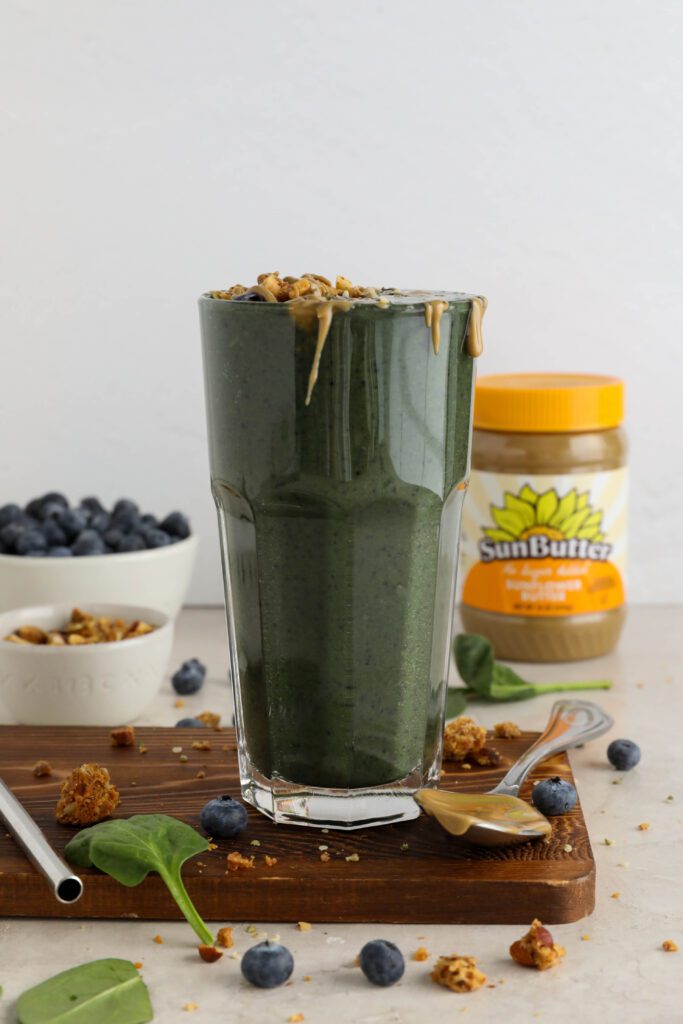 My Go-To Green Smoothie served on wooden board surrounded by blueberries and spinach leaves topped with granola with a spoon with SunButter next to the glass by Flora & Vino