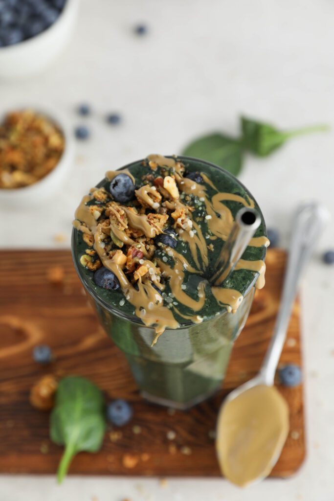 My Go-To Green Smoothie served on wooden board surrounded by blueberries and spinach leaves topped with granola with a spoon with SunButter next to the glass by Flora & Vino