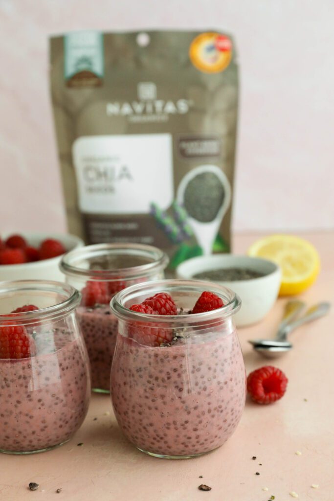 Raspberry Chia Pudding served in mason jars topped with fresh raspberries, cacao nibs, and hemp hearts with two spoons with Navitas Organics package behind by Flora & Vino