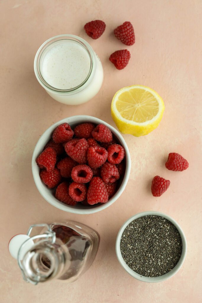 fresh raspberries in a bowl, chia seeds in a bowl, maple syrup in a jar, half of a lemon, and a glass of almond milk on a pink wooden board by Flora & Vino