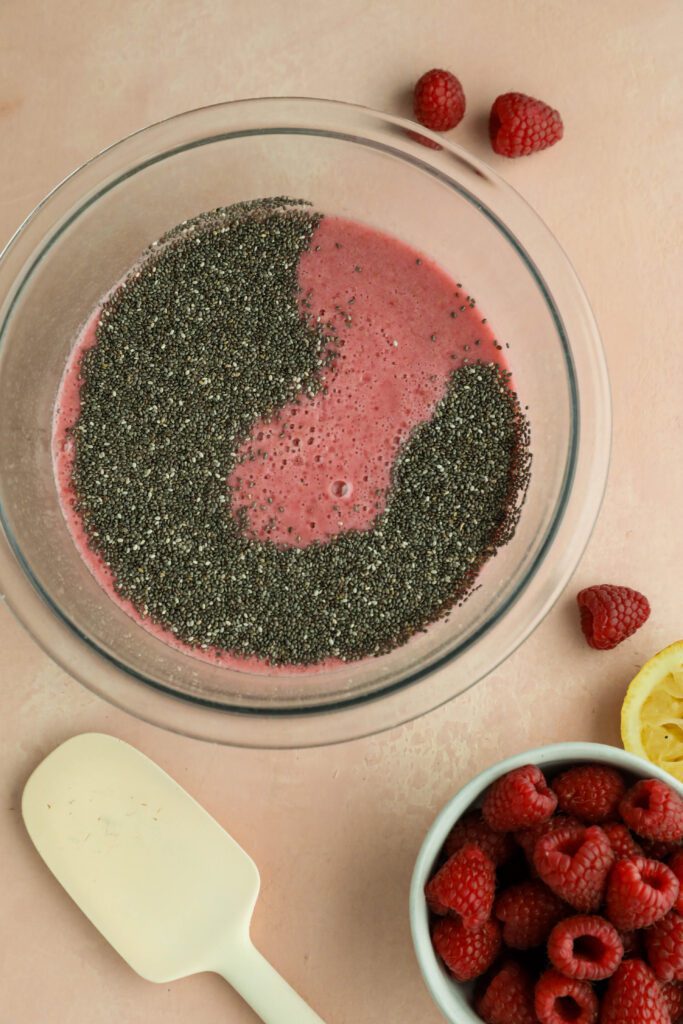 raspberry puree in a glass bowl with chia seeds on top surrounded by fresh raspberries, half of a lemon, and a spatula by Flora & Vino