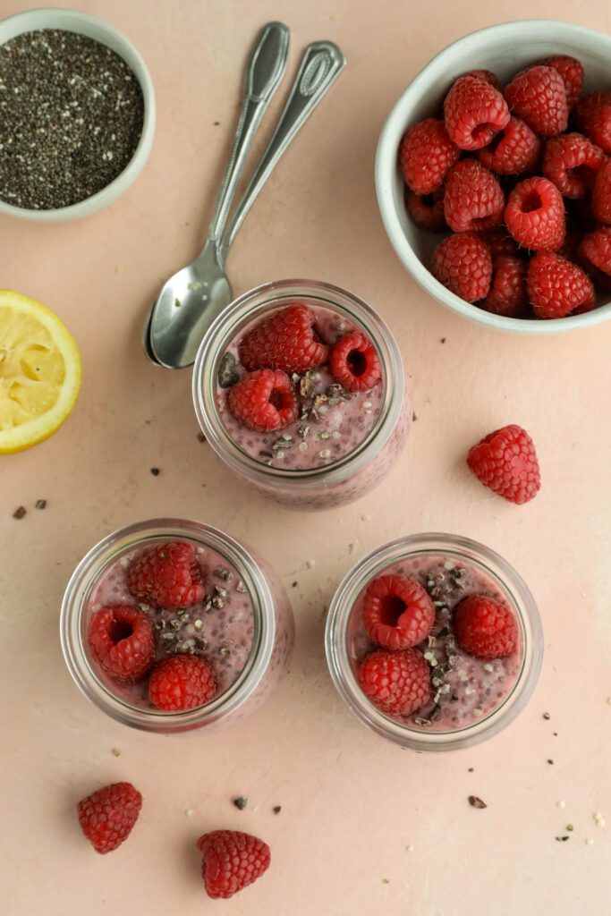 Raspberry Chia Pudding served in mason jars topped with fresh raspberries, cacao nibs, and hemp hearts with two spoons by Flora & Vino