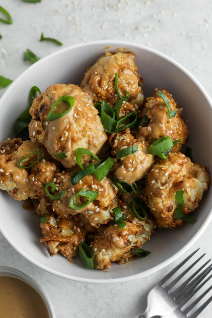 Spicy SunButter Cauliflower Wings served in a bowl and topped with sesame seeds and green onion with two forks and a bowl of SunButter sauce on the side by Flora & Vino