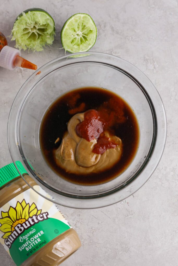 ingredients for Spicy SunButter sauce in glass bowl with sriracha, SunButter, and lime on the side by Flora & Vino