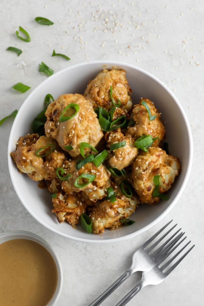 Spicy SunButter Cauliflower Wings served in a bowl and topped with sesame seeds and green onion with two forks and a bowl of SunButter sauce on the side by Flora & Vino