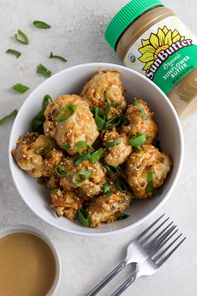 Spicy SunButter Cauliflower Wings served in a bowl and topped with sesame seeds and green onion with two forks and a bowl of SunButter sauce on the side by Flora & Vino