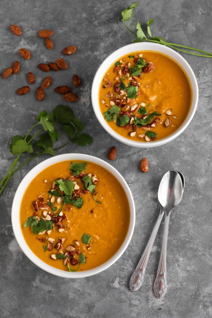 Thai Carrot Sweet Potato Soup served in bowls with two spoons on the side topped with shredded cilantro and Tamari almonds by Flora & Vino