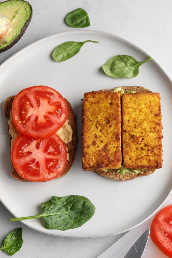 two pieces of toasted sourdough bread on a plate. One half has smashed avocado and breakfast tofu and the other half has hummus and two slices of tomato. The plate is scattered with spinach leaves.
