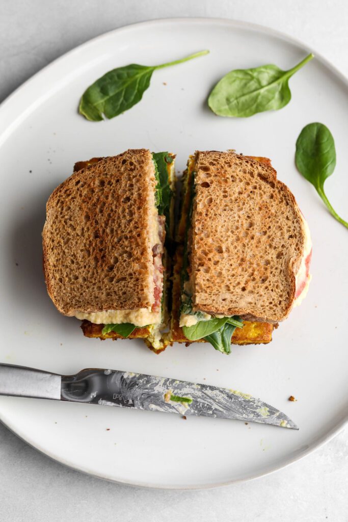 tofu protein breakfast sandwich on plate scattered with spinach and sliced in half with the knife on the side. 