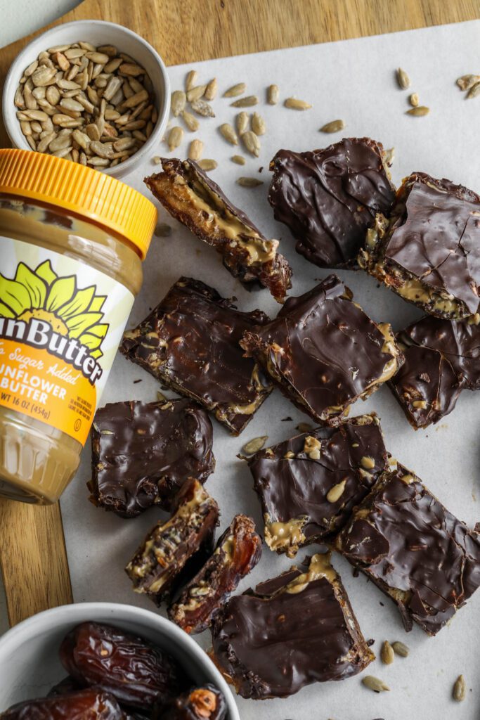 Viral Date Bark (Nut-Free!) cut into squares and served on parchment lined wooden board with bowl of Medjool dates, bowl of sunflower seeds, and spoon of SunButter by Flora & Vino