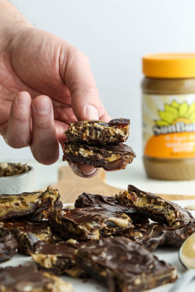 handing holding Viral Date Bark (Nut-Free!) cut into squares and served on parchment lined wooden board with bowl of Medjool dates, bowl of sunflower seeds, and spoon of SunButter by Flora & Vino