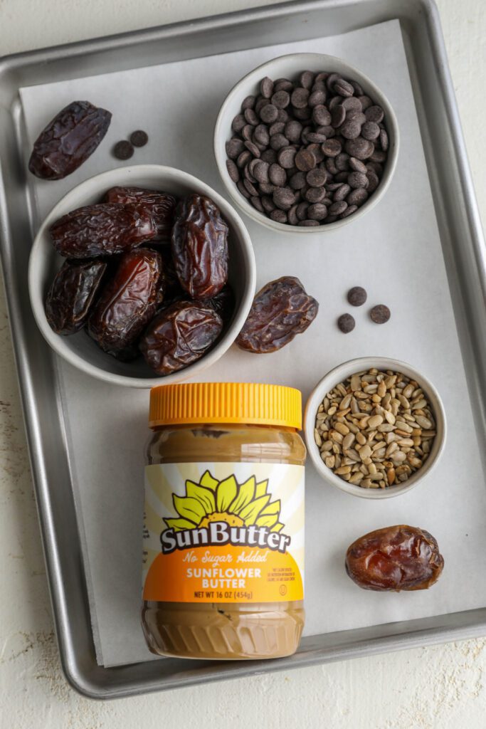jar of No Sugar Added SunButter, bowl of Medjool dates, and bowl of dark chocolate chips, bowl of sunflower seeds on parchment lined baking sheet by Flora & Vino