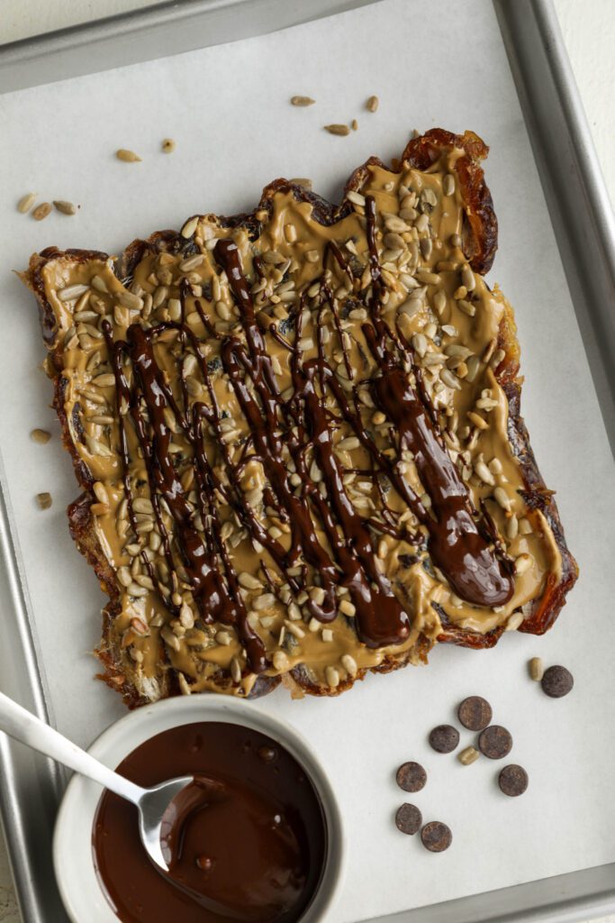 melted dark chocolate drizzled on Medjool date bark with SunButter and sunflower seeds on parchment lined baking sheet with bowl of melted chocolate and spoon by Flora & vino
