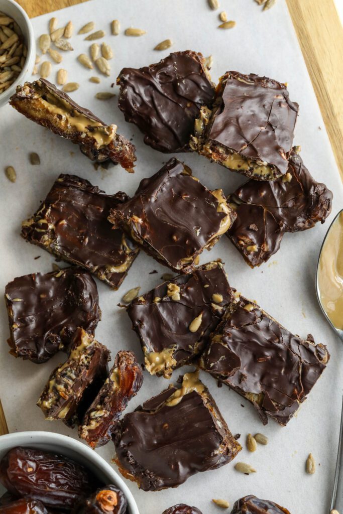 Viral Date Bark (Nut-Free!) cut into squares and served on parchment lined wooden board with bowl of Medjool dates, bowl of sunflower seeds, and spoon of SunButter by Flora & Vino