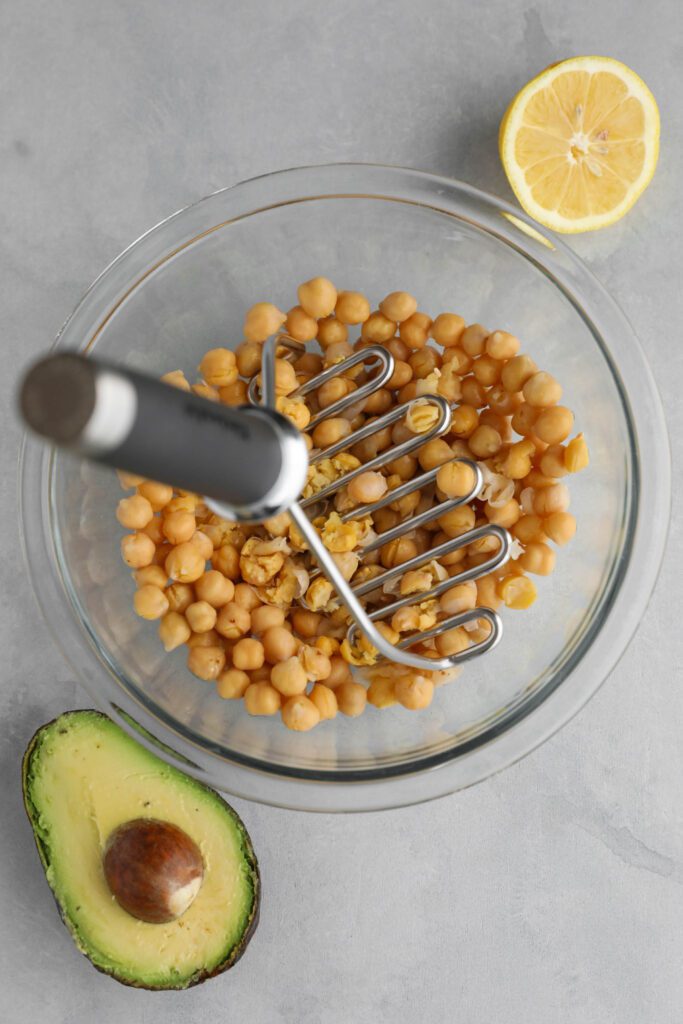 chickpeas in bowl being smashed with potato smasher with lemon and half avocado on the side