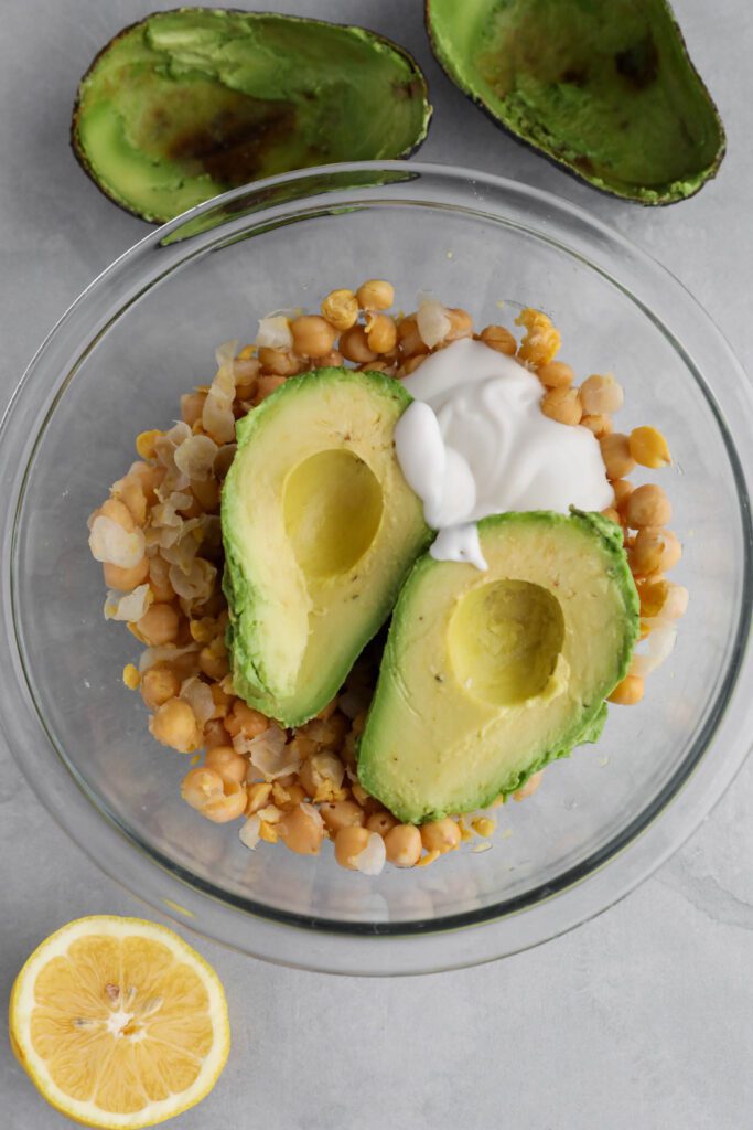 smashed chickpeas in a glass bowl with two avocado halves with avocado shells and half lemon on the side 