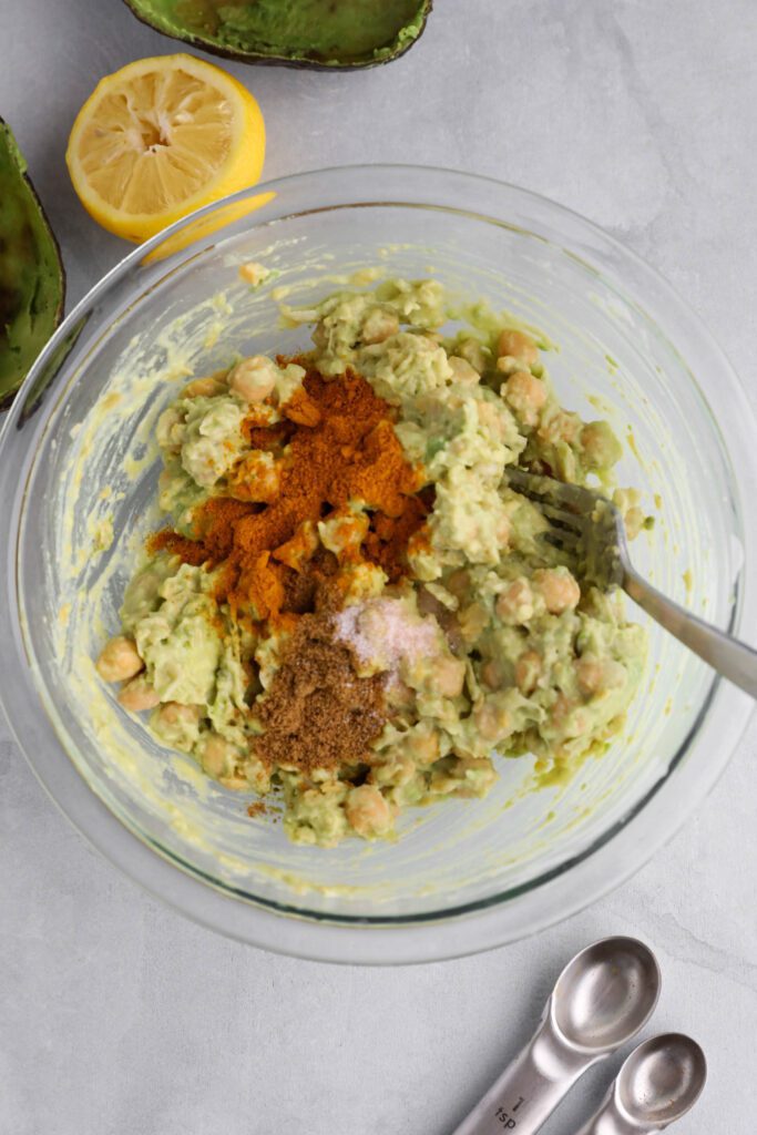 Curried Chickpea Salad being mixed in glass bowl with a fork with lemon and avocado shells on the side