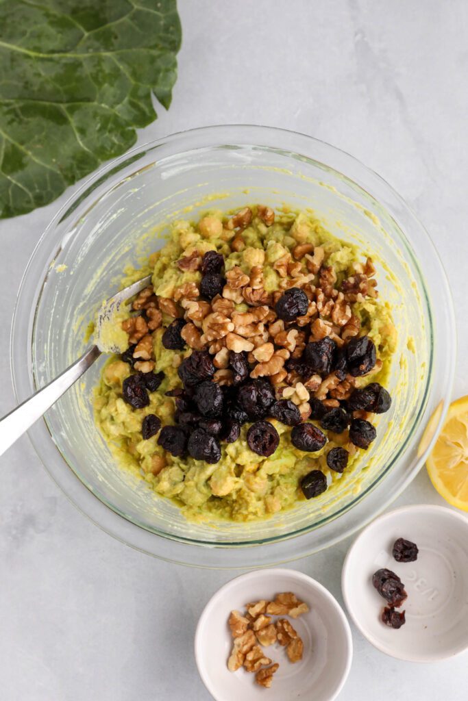 Curried Chickpea Salad mixed in glass bowl with a fork with dried cranberries and crushed walnuts on top with collard leaf on the side 
