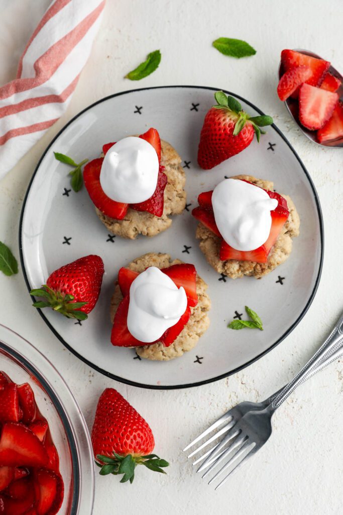 Vegan Cashini Strawberry Shortcakes served on black and white plate with coconut whipped cream and fresh mint with pink and white towel, spoon of marinated strawberries, and two forks 