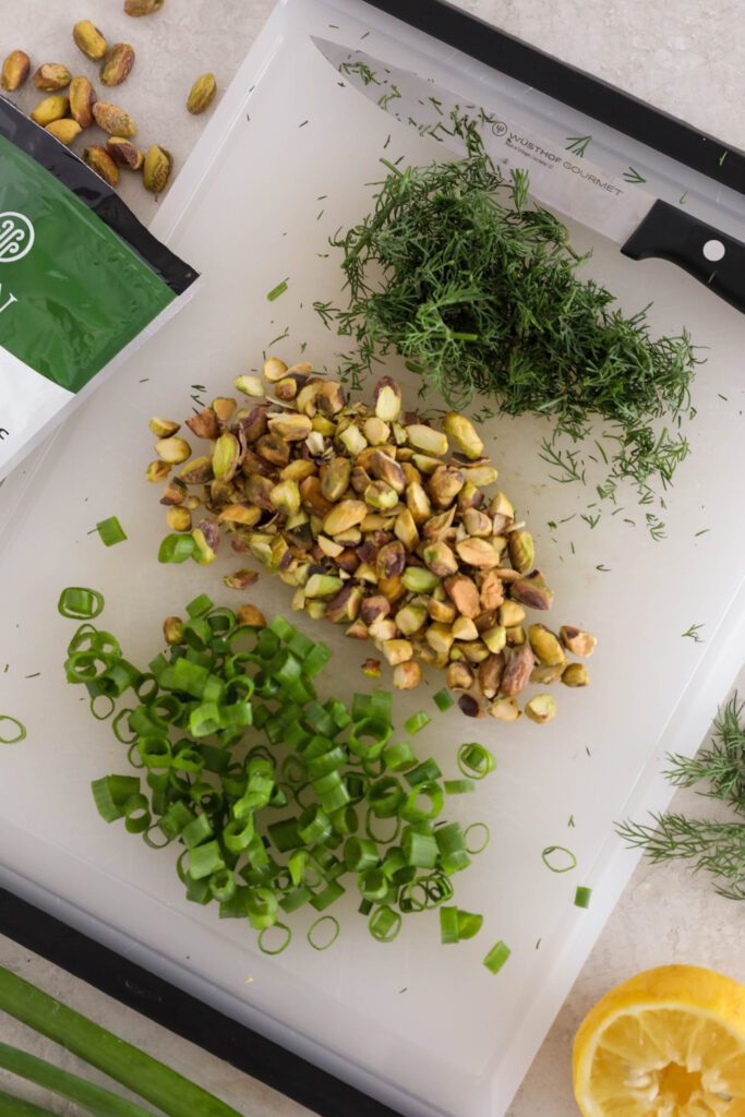 fresh dill, chopped pistachios, and sliced green onion on cutting board with a knife with dill, lemon, green onion, and pistachios on the side