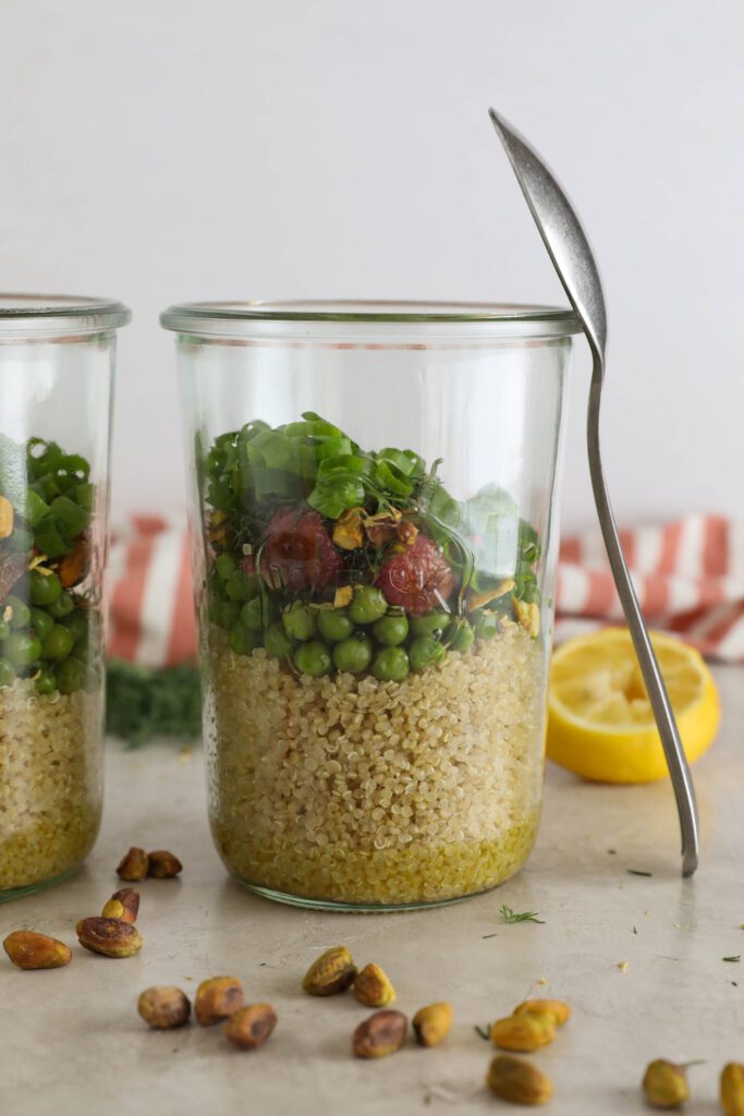two Spring Quinoa Salad Jars with spoon balanced on one with half lemon and scattered pistachios in front of pink and white striped towel