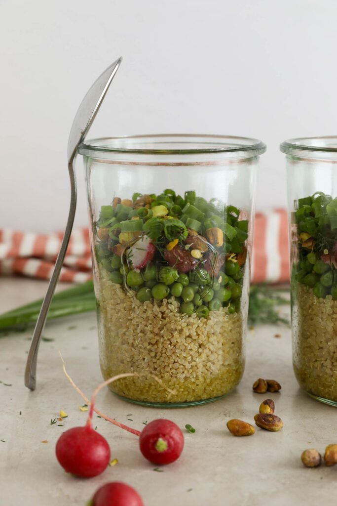 two Spring Quinoa Salad Jars with spoon balanced on one with half lemon and scattered pistachios in front of pink and white striped towel
