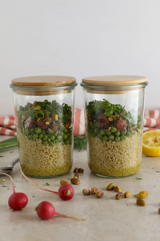 two Spring Quinoa Salad Jars with spoon balanced on one with half lemon and scattered pistachios in front of pink and white striped towel