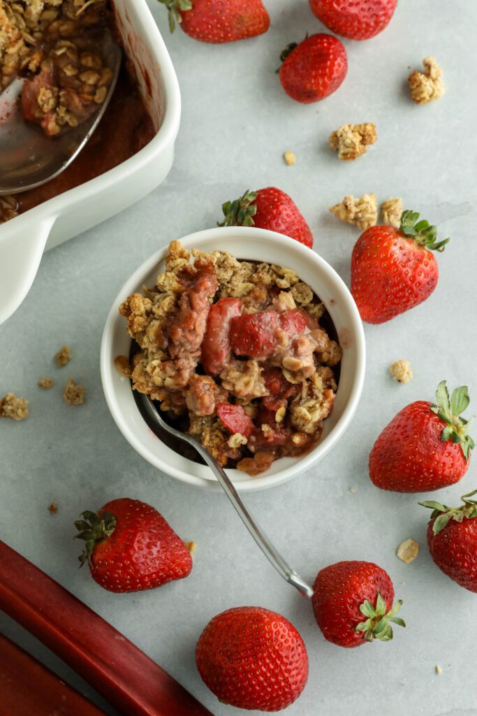 Vegan Strawberry Rhubarb Crumble served in ramekin and spoon and crumble in square baking dish with spoon surrounded by strawberries and rhubarb stalks