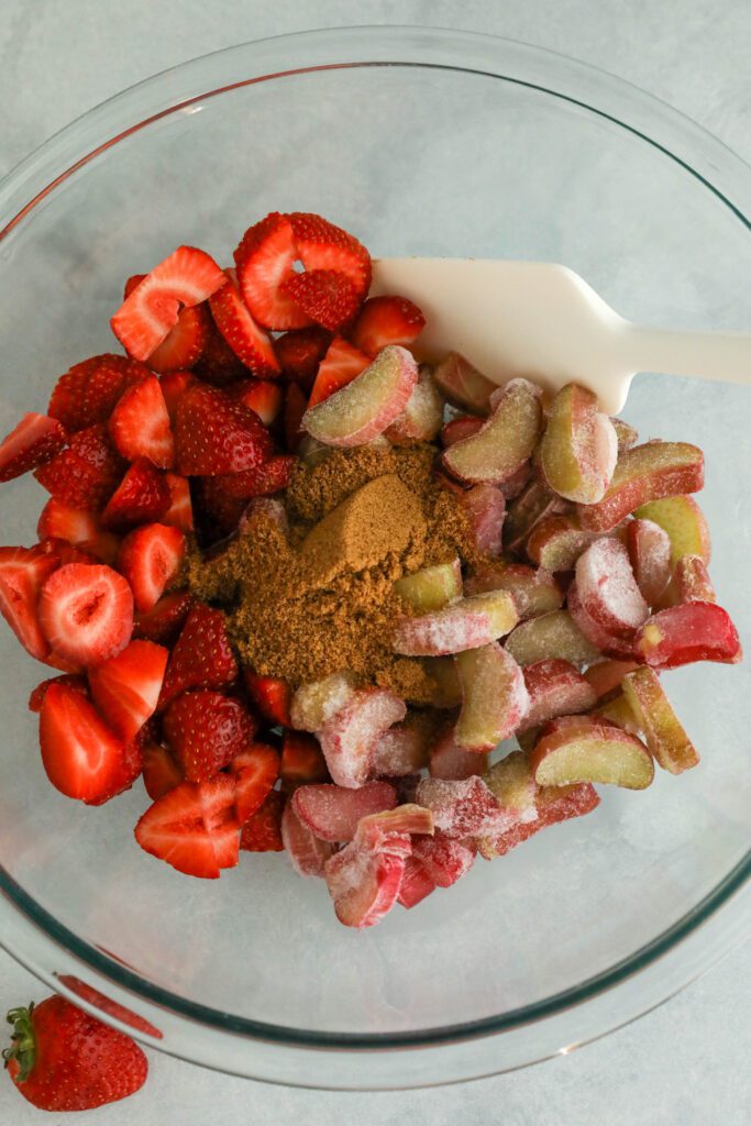 fresh sliced strawberries and frozen rhubarb in glass bowl with coconut sugar and spatula 