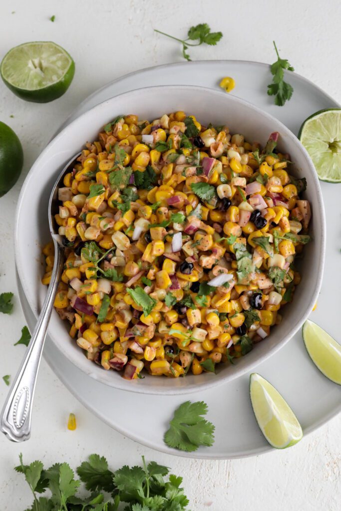 Vegan Elote Corn Salad served in bowl with spoon surrounded by cilantro and limes 