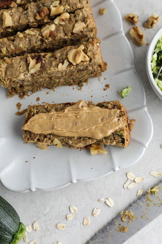 Vegan Zucchini Bread sliced on serving tray with zucchini and walnuts on side. One piece smeared with nut butter