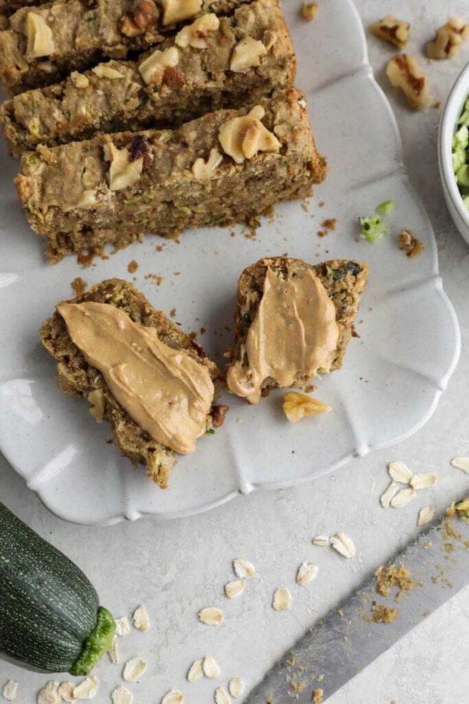 Vegan Zucchini Bread sliced on serving tray with zucchini and walnuts on side. One piece smeared with nut butter