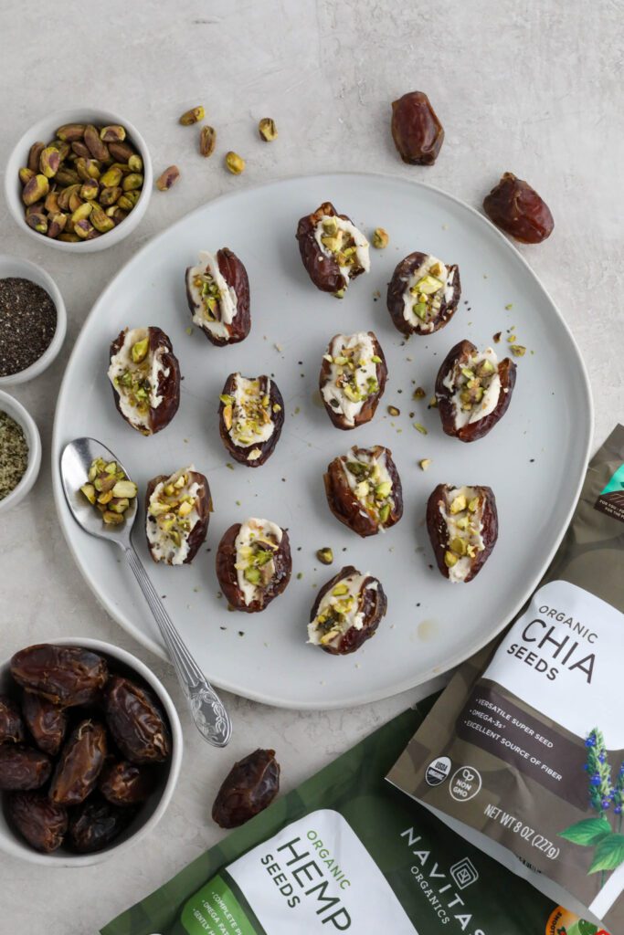 Dates stuffed with cream cheese and topped with pistachios, hemp hearts, and chia seeds on white plate