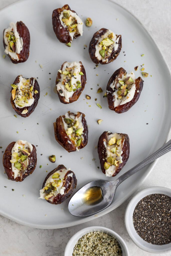 Dates stuffed with cream cheese and topped with pistachios, hemp hearts, and chia seeds on white plate
