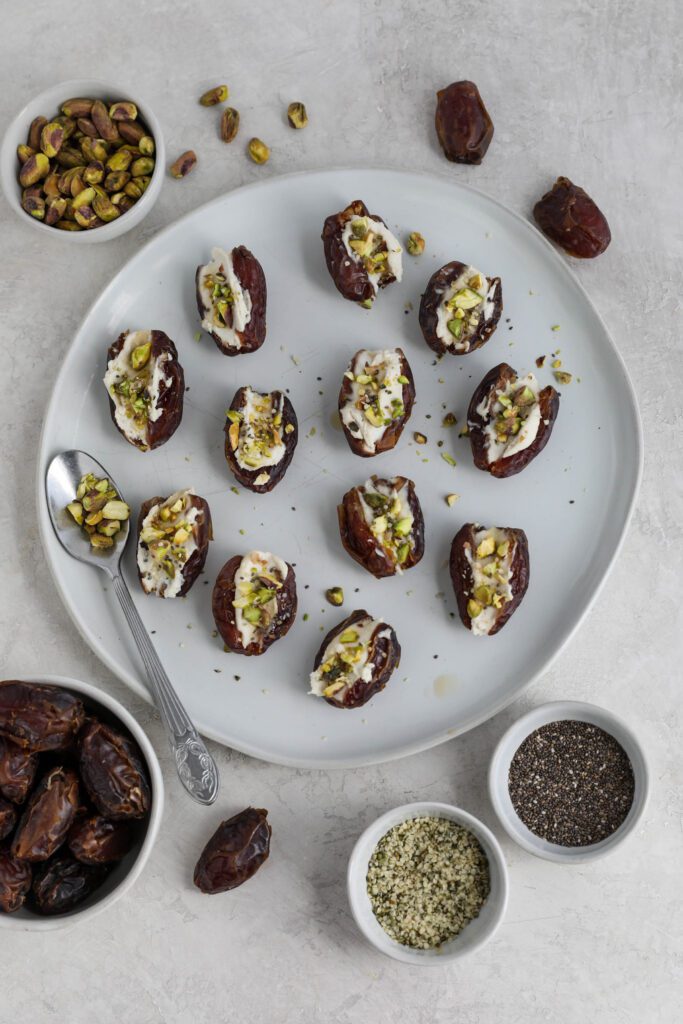 Dates stuffed with cream cheese and topped with pistachios, hemp hearts, and chia seeds on white plate