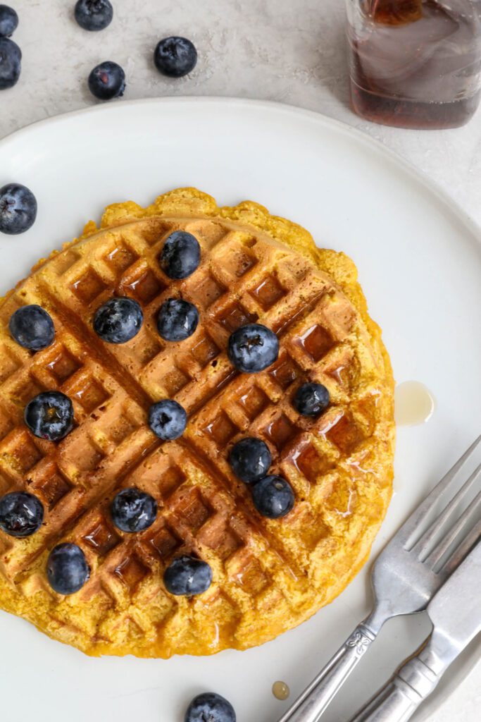 Sweet Potato Waffles served on a plate topped with maple syrup and blueberries with fork and knife