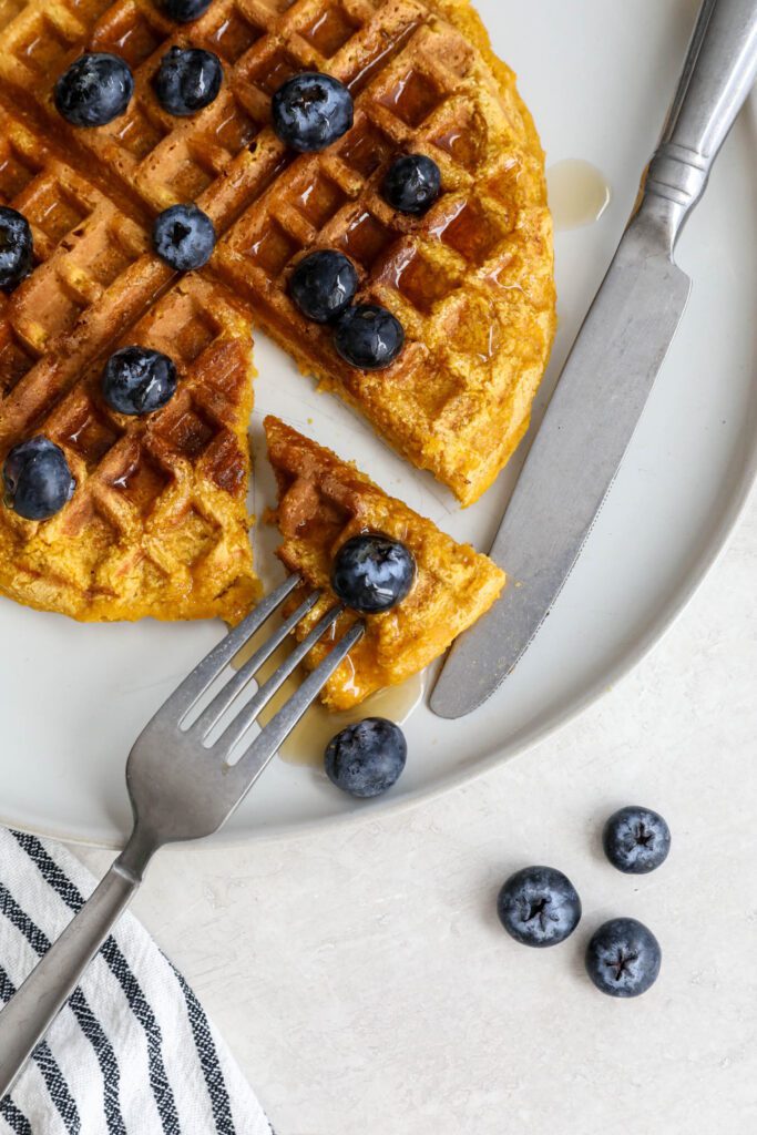 Sweet Potato Waffles served on a plate topped with maple syrup and blueberries with a triangle cut out with fork and knife