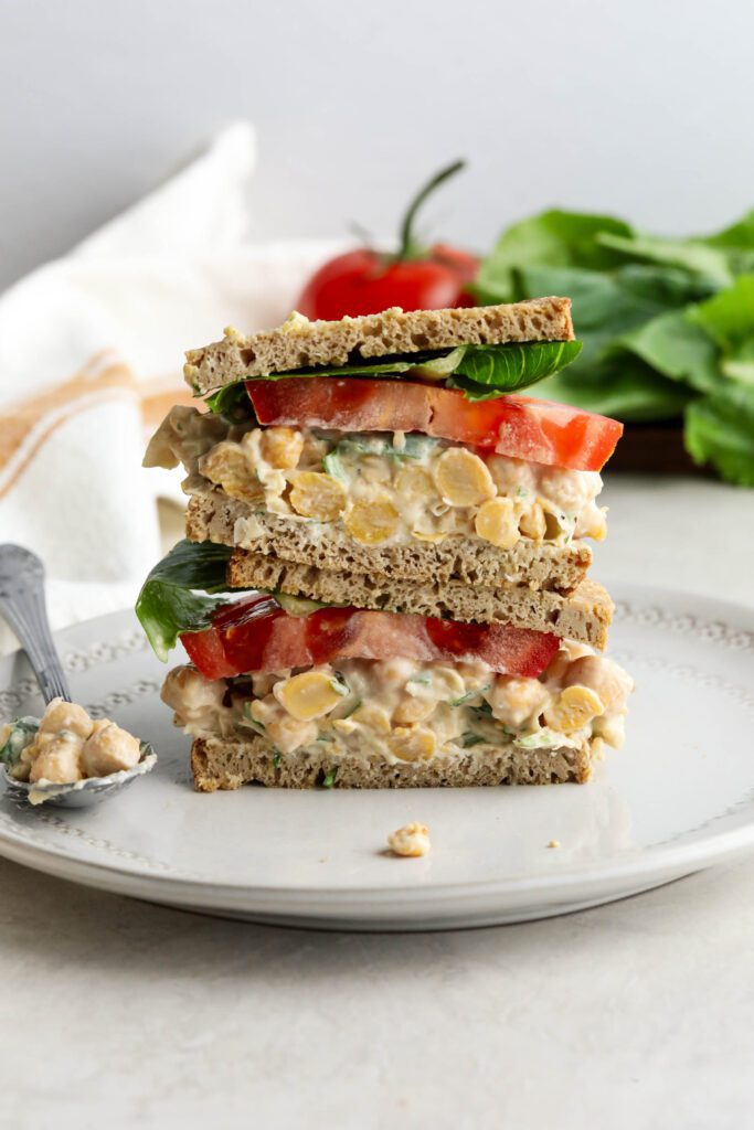 Vegan Chickpea Salad Sandwich stacked on plate with lettuce and tomato in background