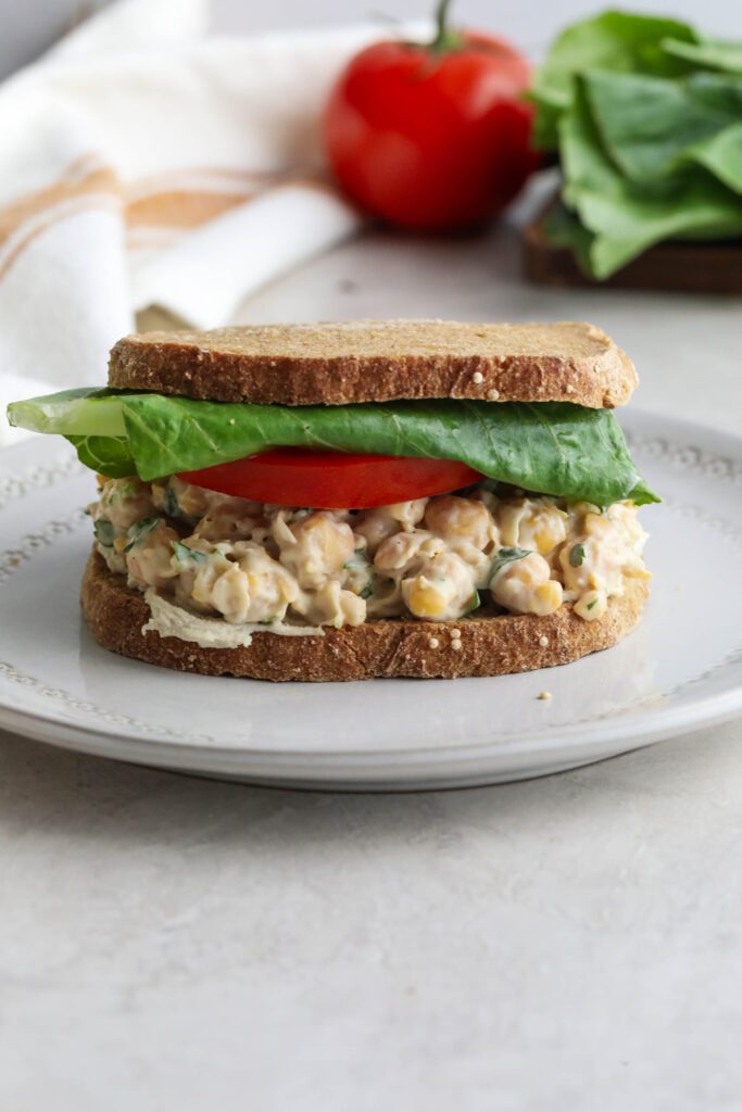 Vegan Chickpea Salad Sandwich stacked on plate with lettuce and tomato in background