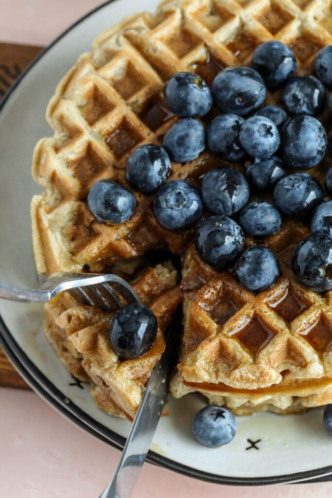 Vegan Applesauce Waffles stacked on plate with blueberries drizzled with maple syrup with fork and knife cutting wedge out