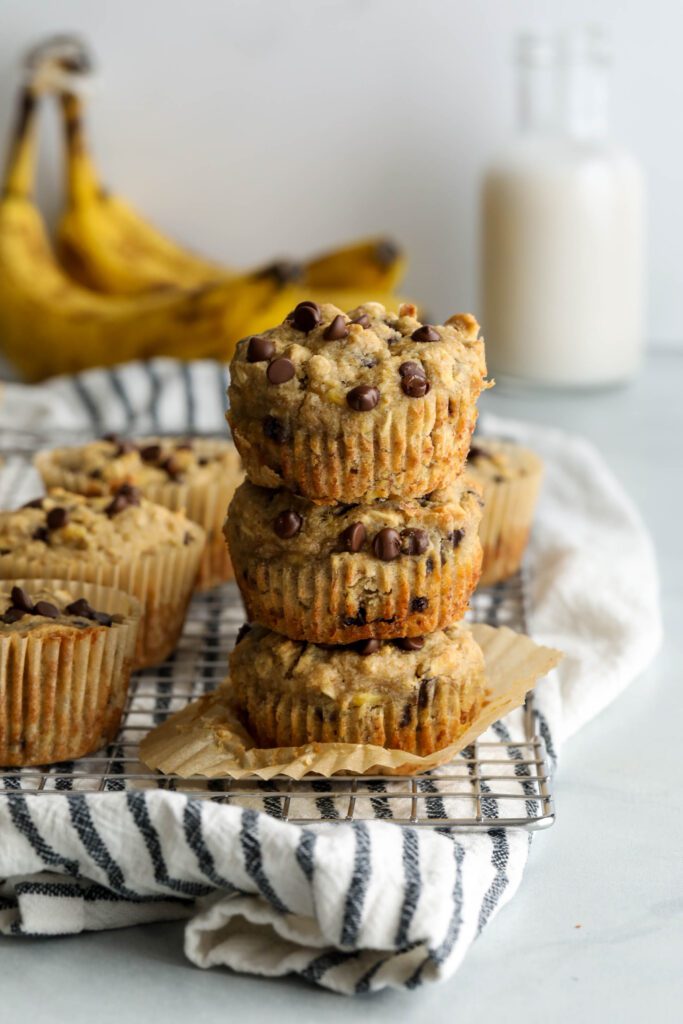 Banana Oatmeal Chocolate Chips Muffins stacked on cooling rack with blue and white striped towel and bananas and milk jar in background