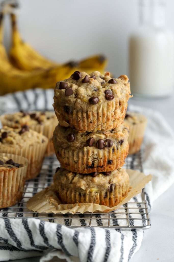 Banana Oatmeal Chocolate Chips Muffins stacked on cooling rack with blue and white striped towel and bananas and milk jar in background