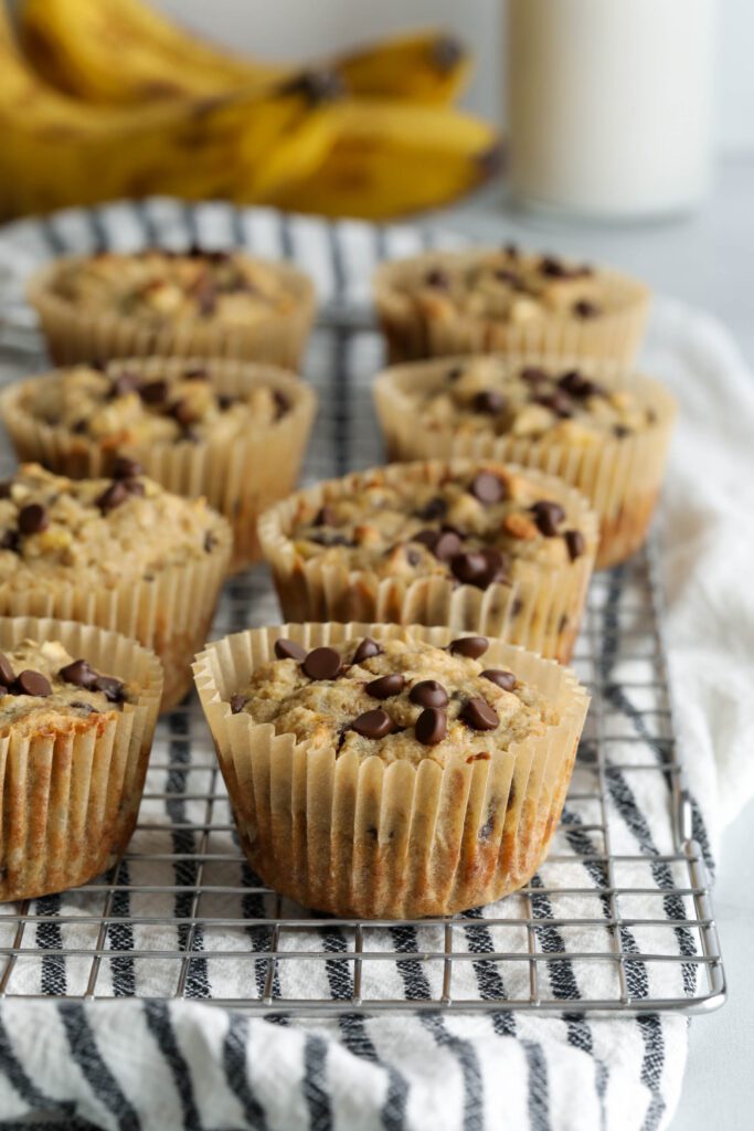Banana Oatmeal Chocolate Chips Muffins stacked on cooling rack with blue and white striped towel and bananas and milk jar in background