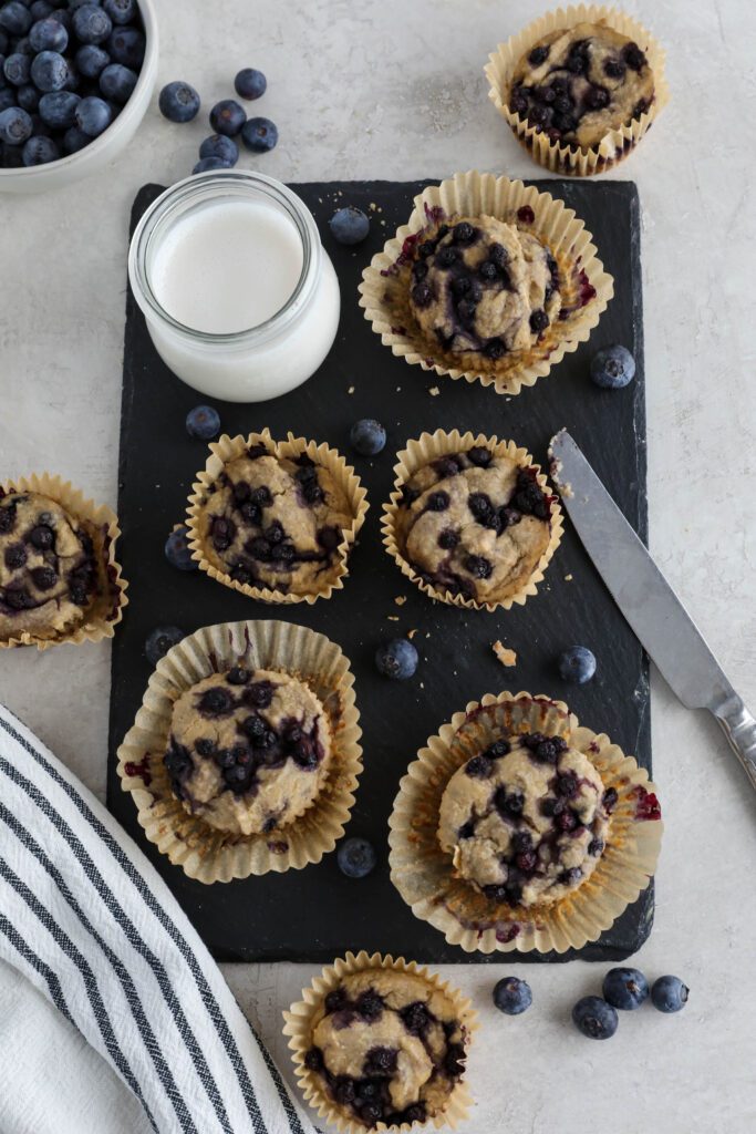 Vegan Blueberry Muffins served on black rectangle with glass of milk, knife, and blueberries 